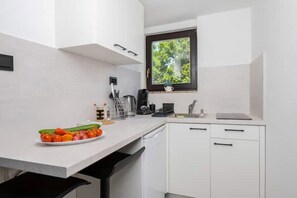 A compact kitchen with white cabinets, a window with a view of greenery, a sink, and a countertop with a plate of vegetables. - Luxury Seaview Apartment (Zagorje)