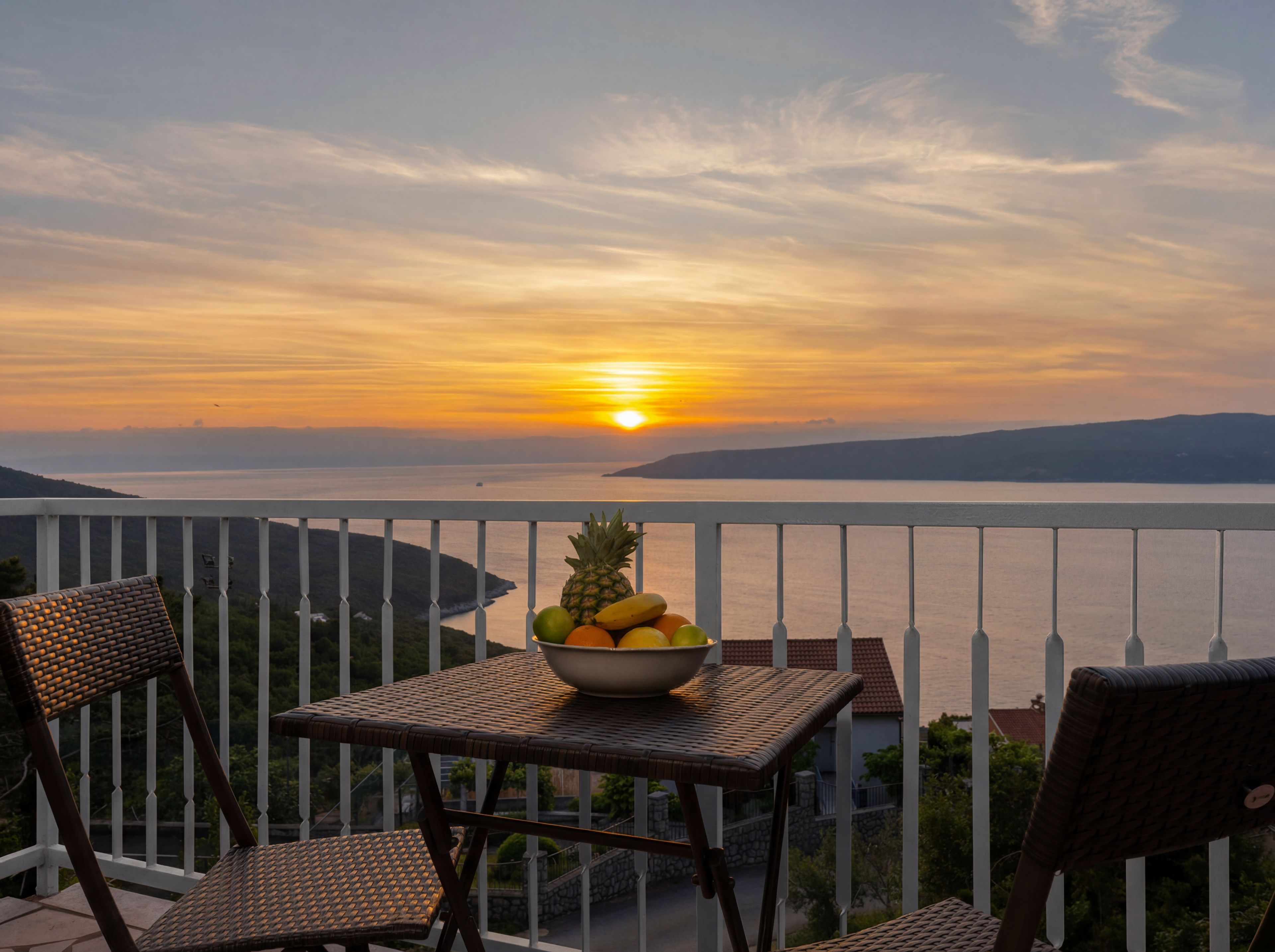 Een balkon met rieten meubilair, een fruitschaal en uitzicht op het water bij zonsondergang.