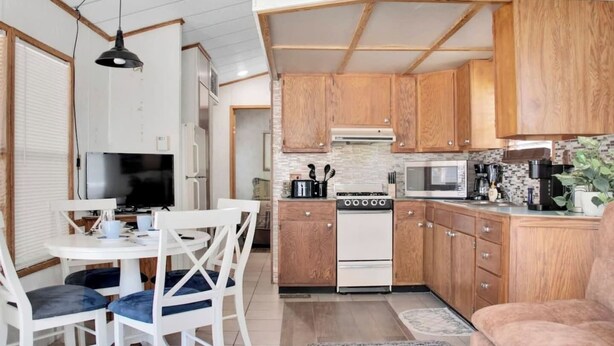 Una cocina funcional con armarios de madera, estufa, microondas y un comedor con televisor y sillas.