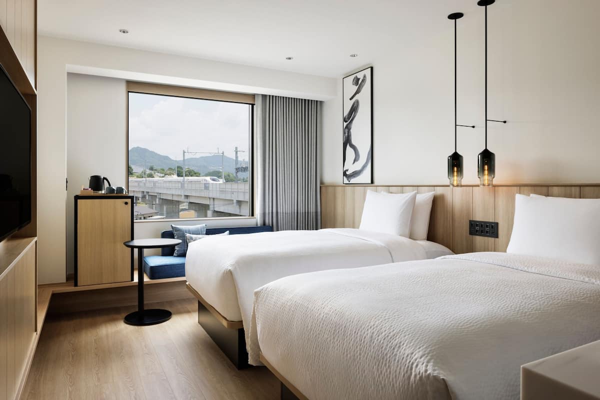 Ein Hotelzimmer mit zwei Betten, einem Fernseher, einem kleinen Tisch und einem Fenster mit Blick auf eine Brücke und Berge.