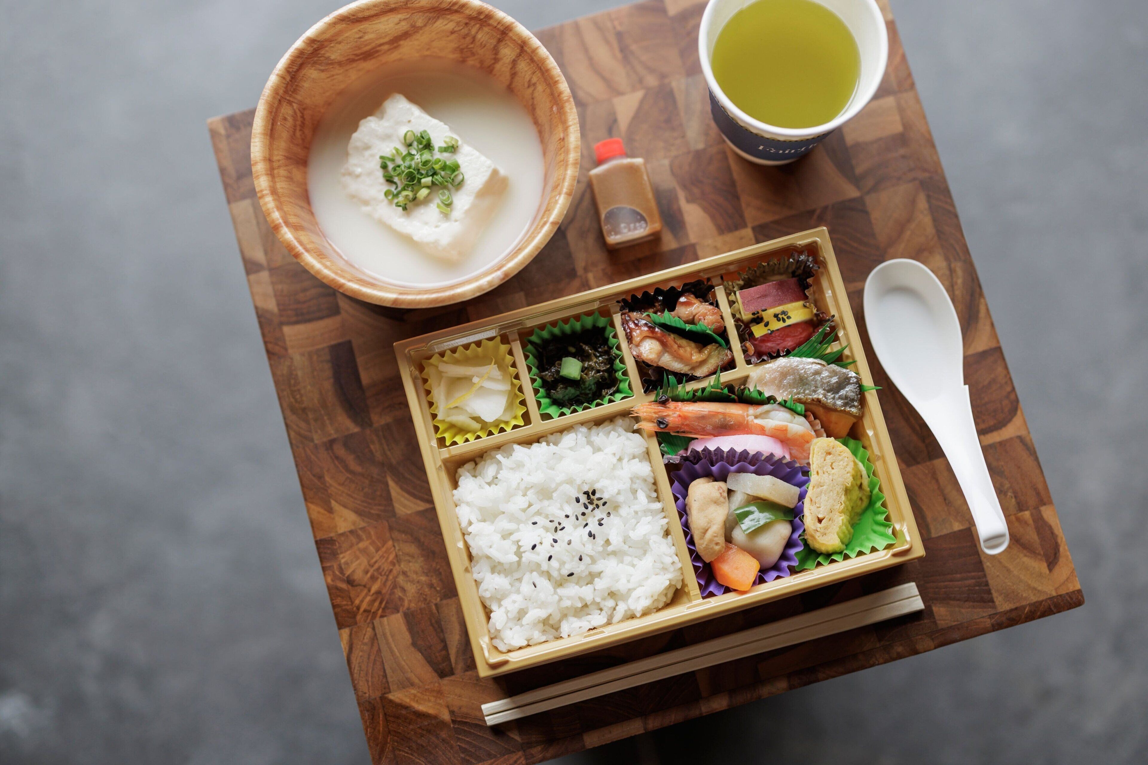 A traditional Japanese meal with rice, fish, and vegetables, served on a wooden tray with chopsticks and a spoon.