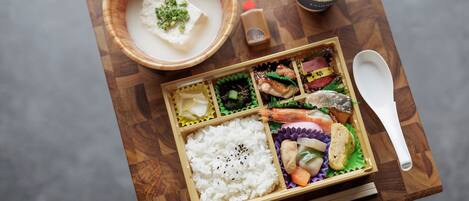 Comida tradicional japonesa con arroz, pescado y verduras, servida en una bandeja de madera con palillos y una cuchara.
