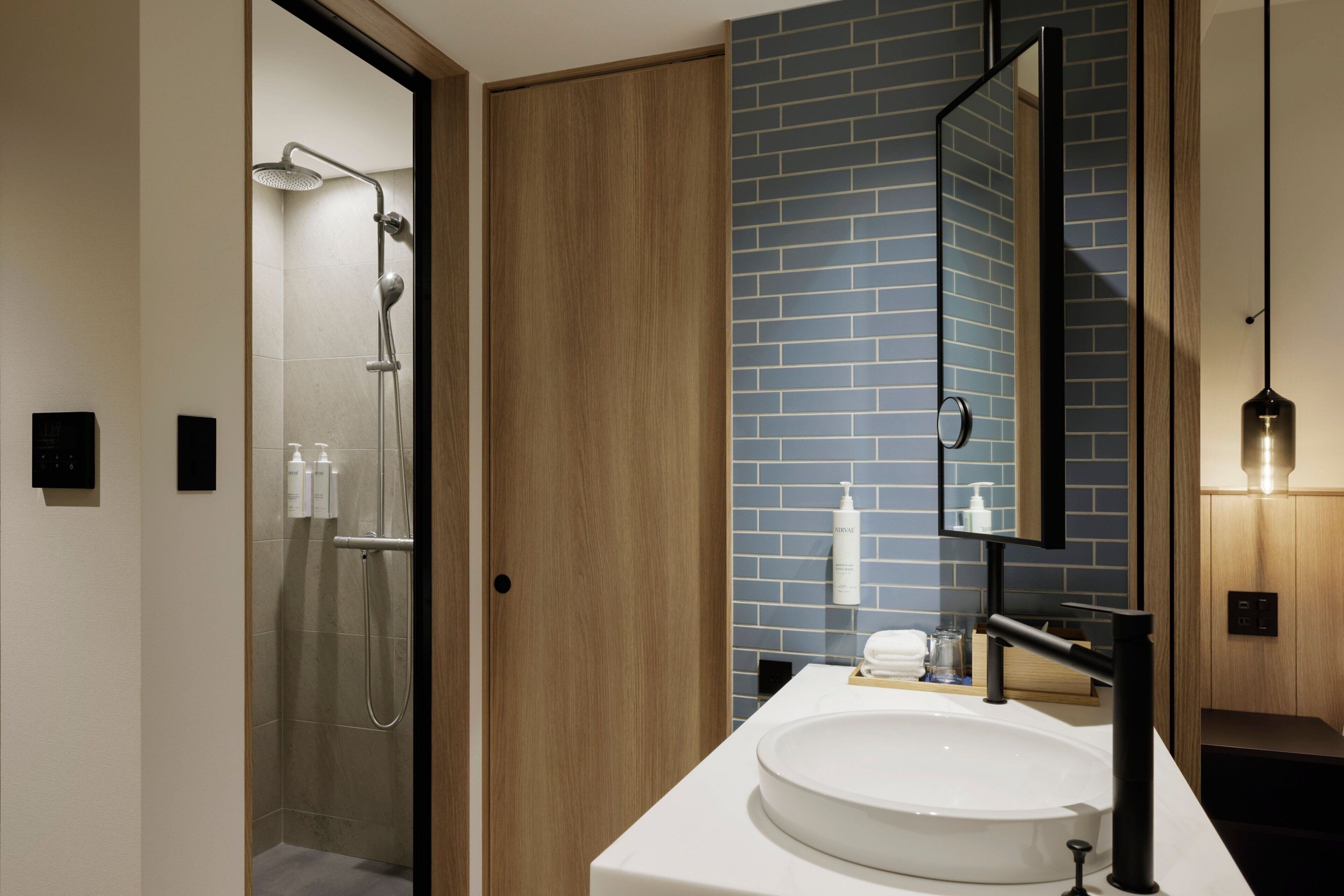 Un baño moderno con ducha de vidrio, puerta de madera, pared con azulejos azules, lavamanos blanco y un espejo.