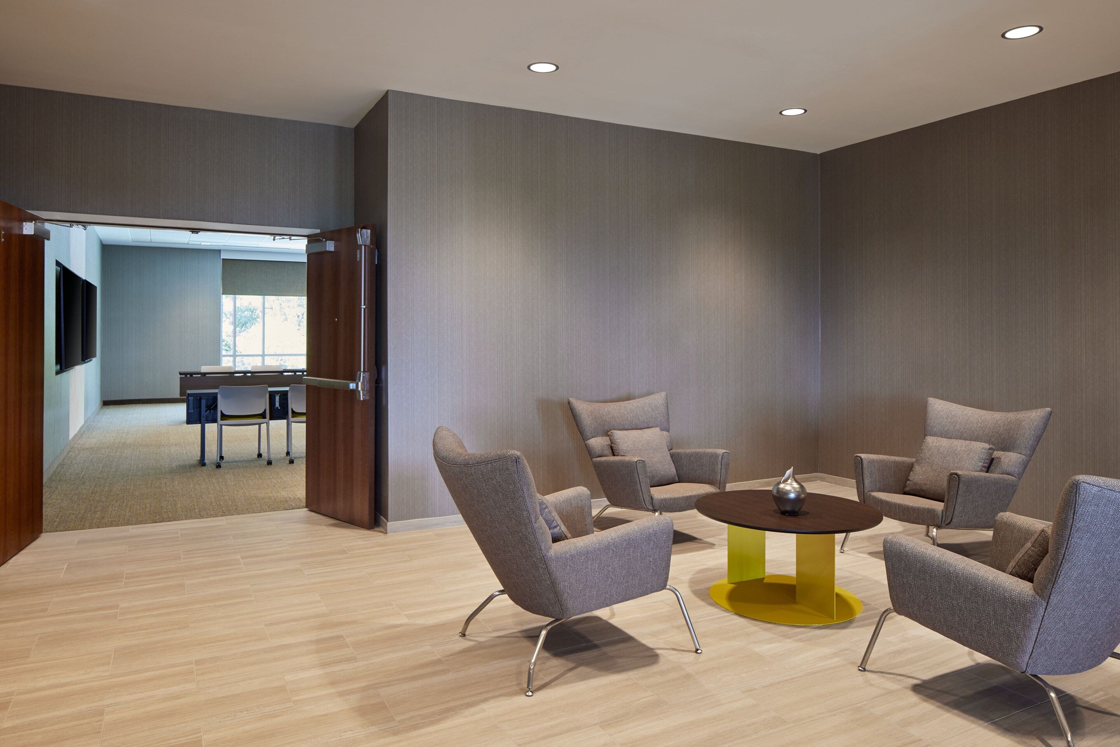 A modern conference room with four grey upholstered chairs around a yellow and brown table, wooden flooring, and a large window in the background.