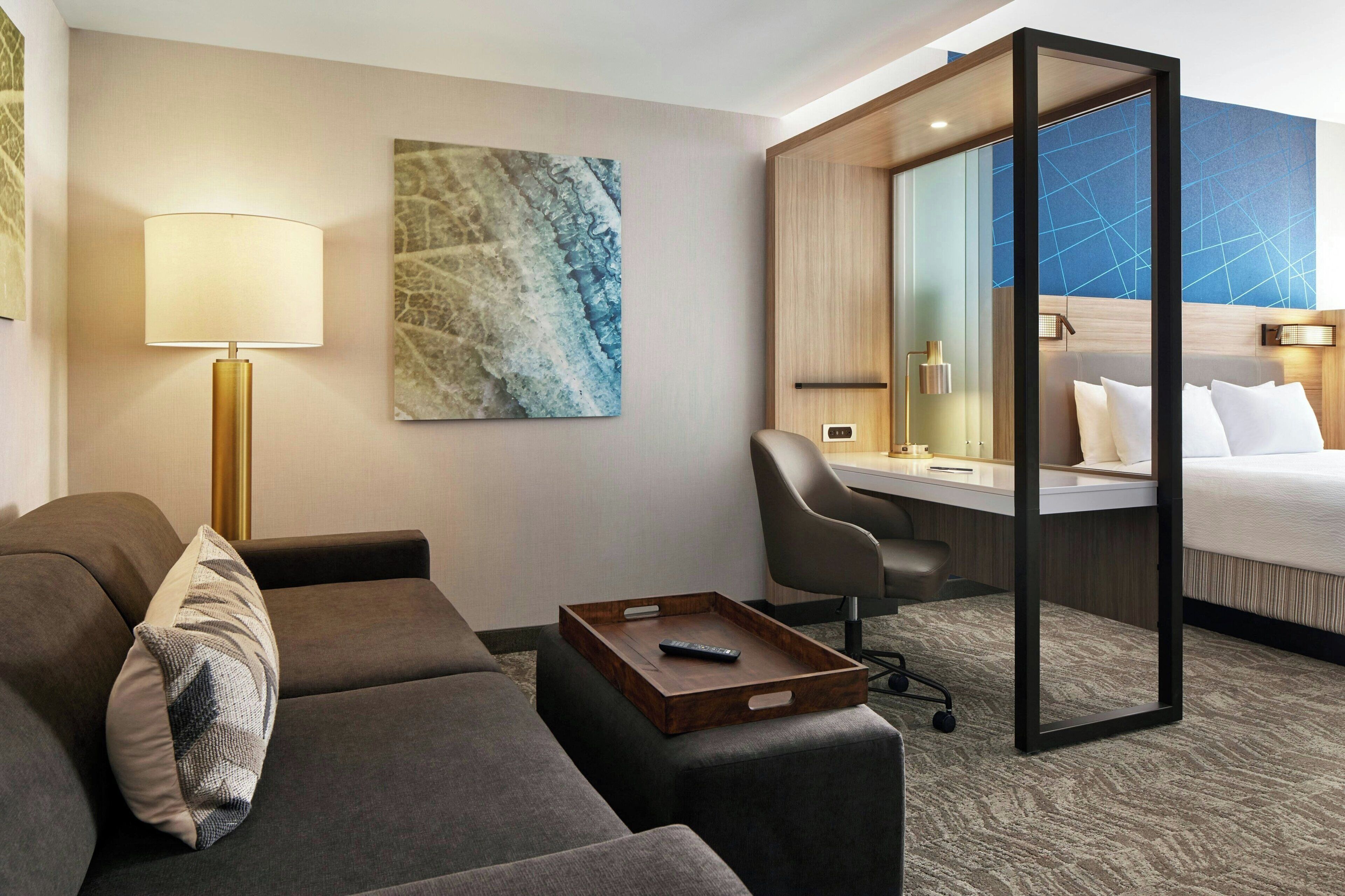 A hotel room with a sofa, a desk, a bed, a lamp, and a painting on the wall.