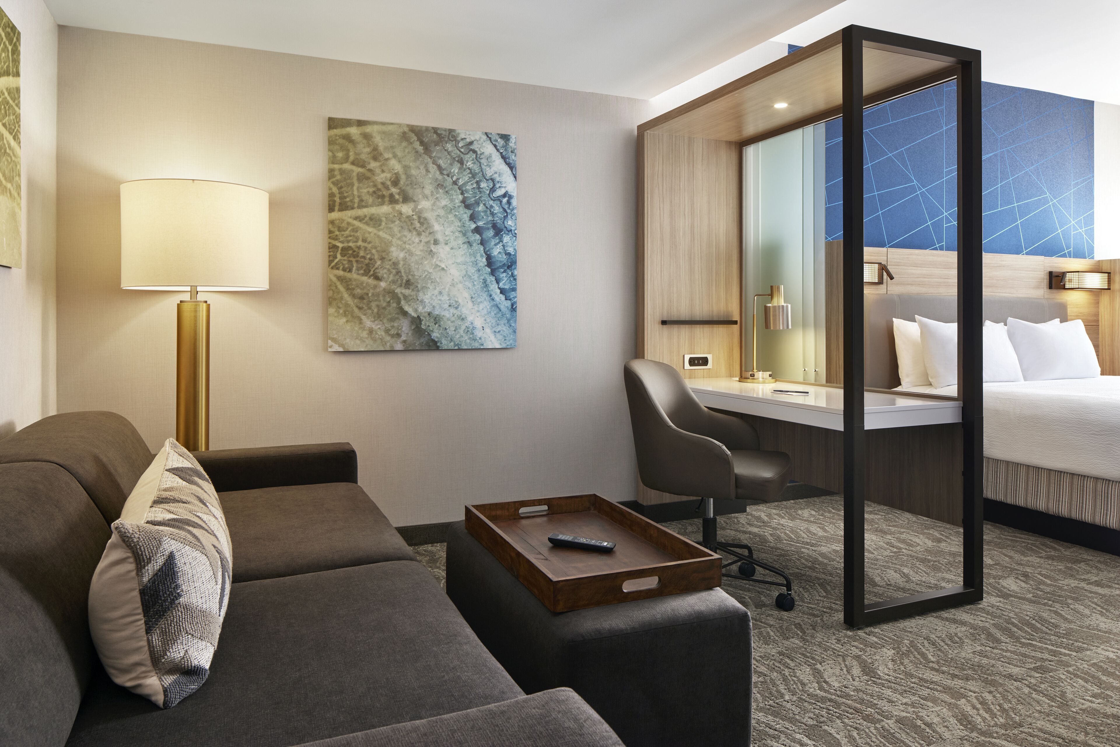 A hotel room with a sofa, a desk, a bed, a lamp, and a painting on the wall.