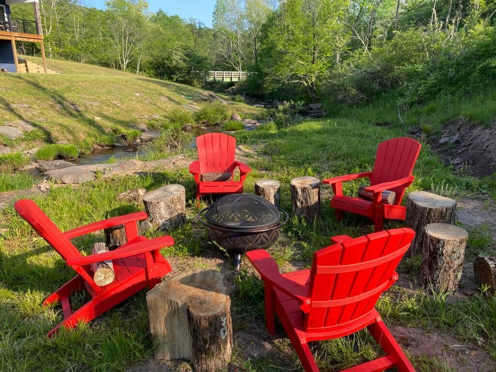 Un espace extérieur aménagé avec des sièges disposés en cercle autour d’un foyer, composé de chaises Adirondack rouges. L’ensemble est situé dans une zone herbeuse, avec des arbres et un ruisseau en arrière-plan.