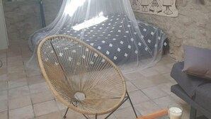 A cozy room with a bed covered by a mosquito net, a wicker chair, a sofa, and a table with a glass. - Joli Mazet Indépendant à Louer (robiac-rochessadoule)