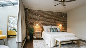 Une chambre avec un mur en briques, un lit avec une literie blanche, un banc, une table de chevet avec une lampe et une grande fenêtre.