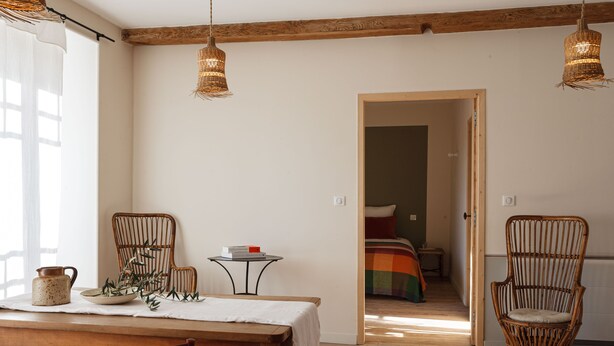 A room with a wooden dining table, wicker chairs, a small round table with a book, and a bed with a colorful blanket in the background.