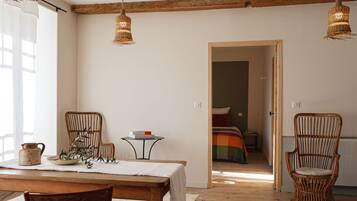 Une pièce avec une table à manger en bois, des chaises en osier, une petite table ronde sur laquelle repose un livre, et un lit avec une couverture colorée en arrière-plan.