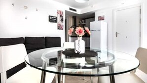 A modern living room with a glass dining table, a black sofa, and a white chair. There is a vase with flowers on the table.