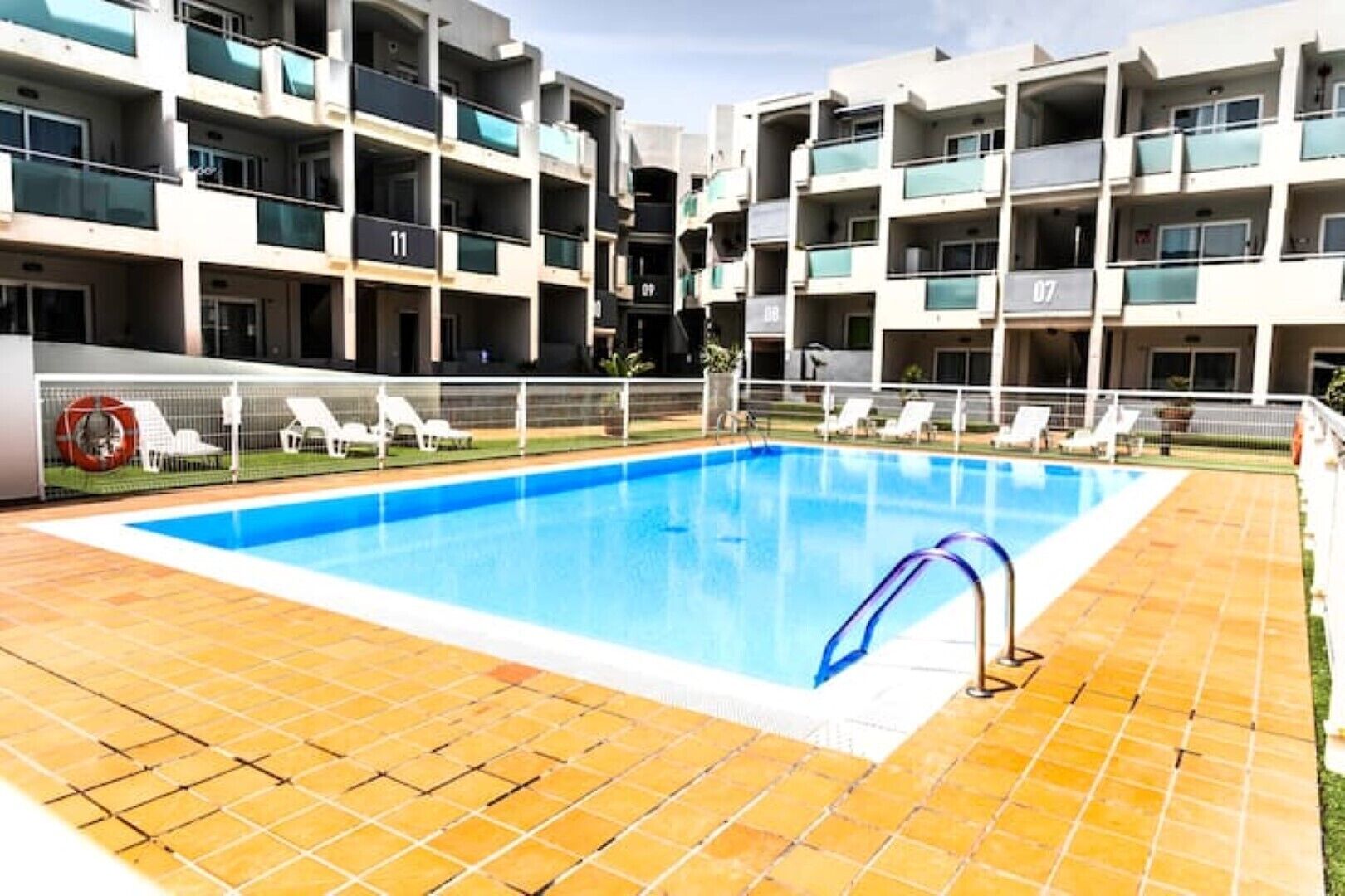 A swimming pool surrounded by a tiled deck, with multiple balconies and lounge chairs in the background.