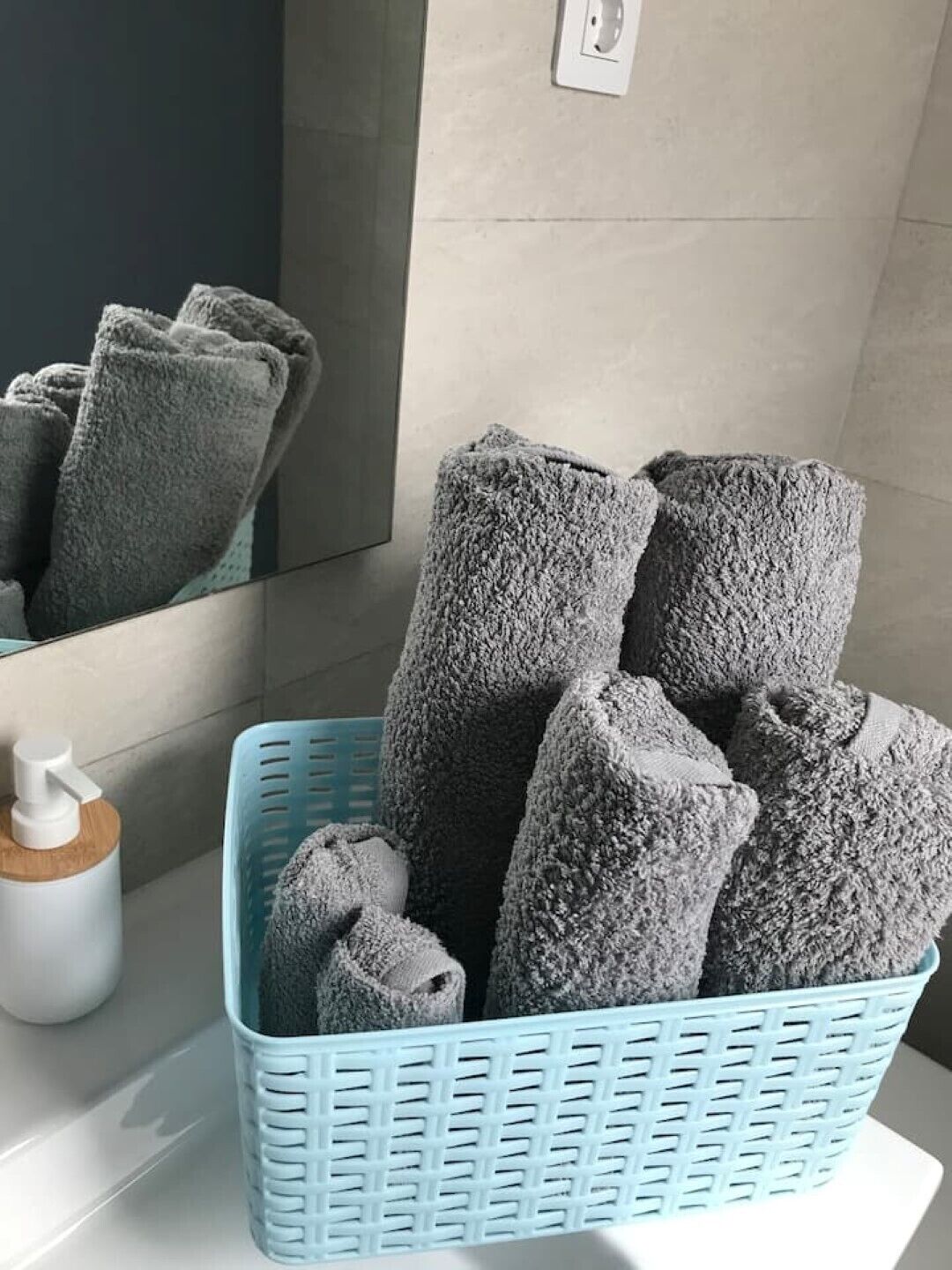 Un baño con una cesta azul que contiene toallas enrolladas, un dispensador de jabón y un espejo que refleja las toallas.