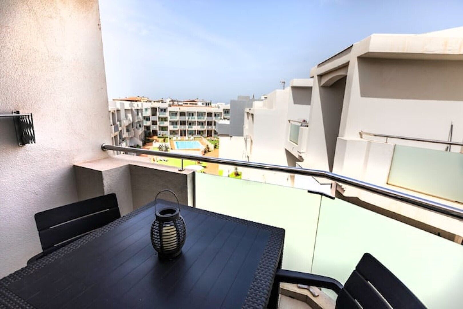 A balcony with a table, chairs, and a decorative lantern. A pool and buildings are visible in the background.