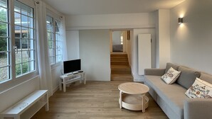 Una sala moderna con un ventanal grande, un televisor de pantalla plana, una mesita redonda de madera, un sofá color beige con cojines estampados y una escalera que lleva a otra habitación.