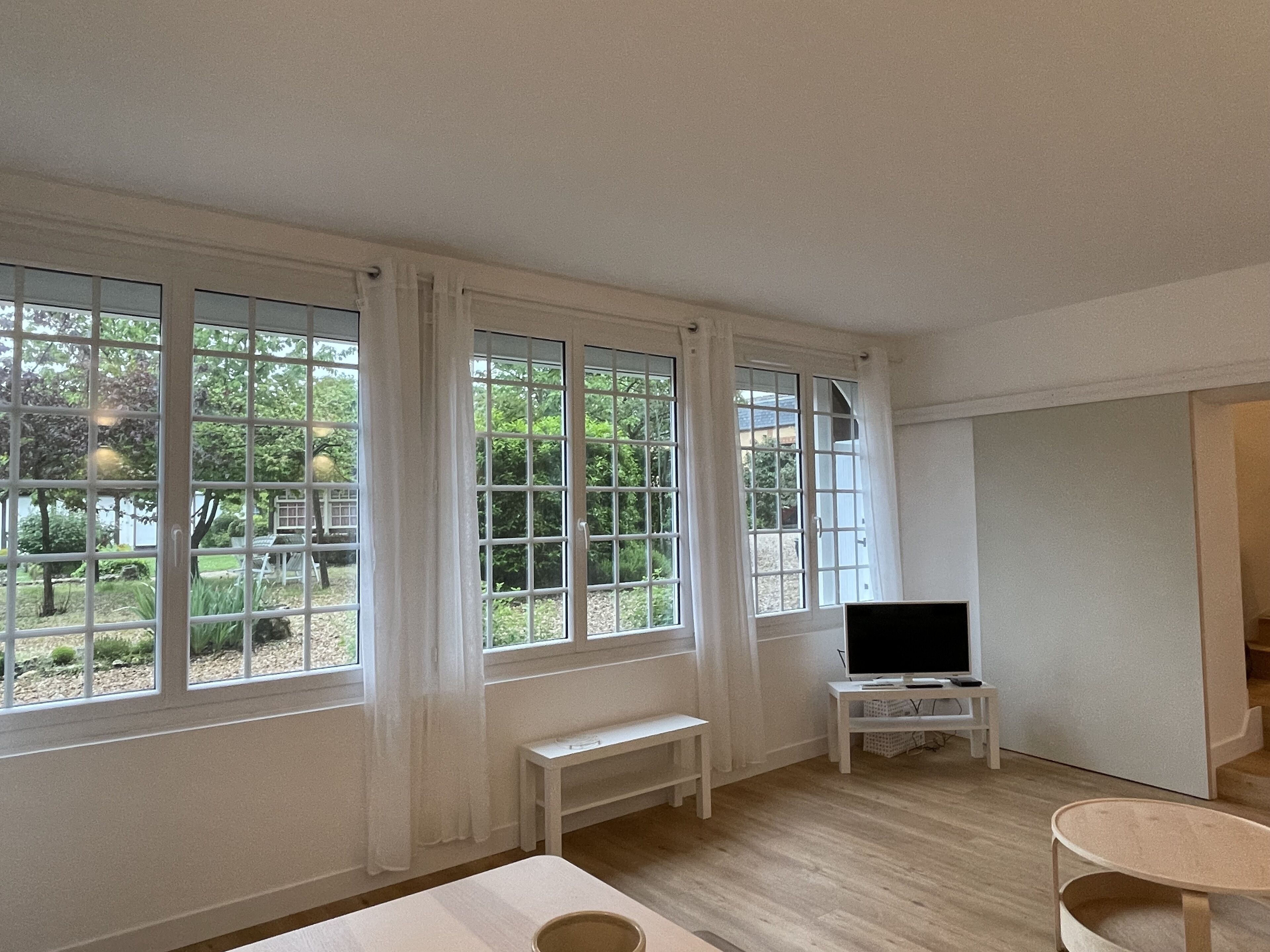 A room with large windows, a television on a stand, a small bench, and a round table.