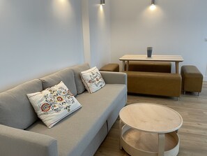 A modern living room with a grey sofa, two patterned cushions, a round wooden coffee table, and a brown sofa set.