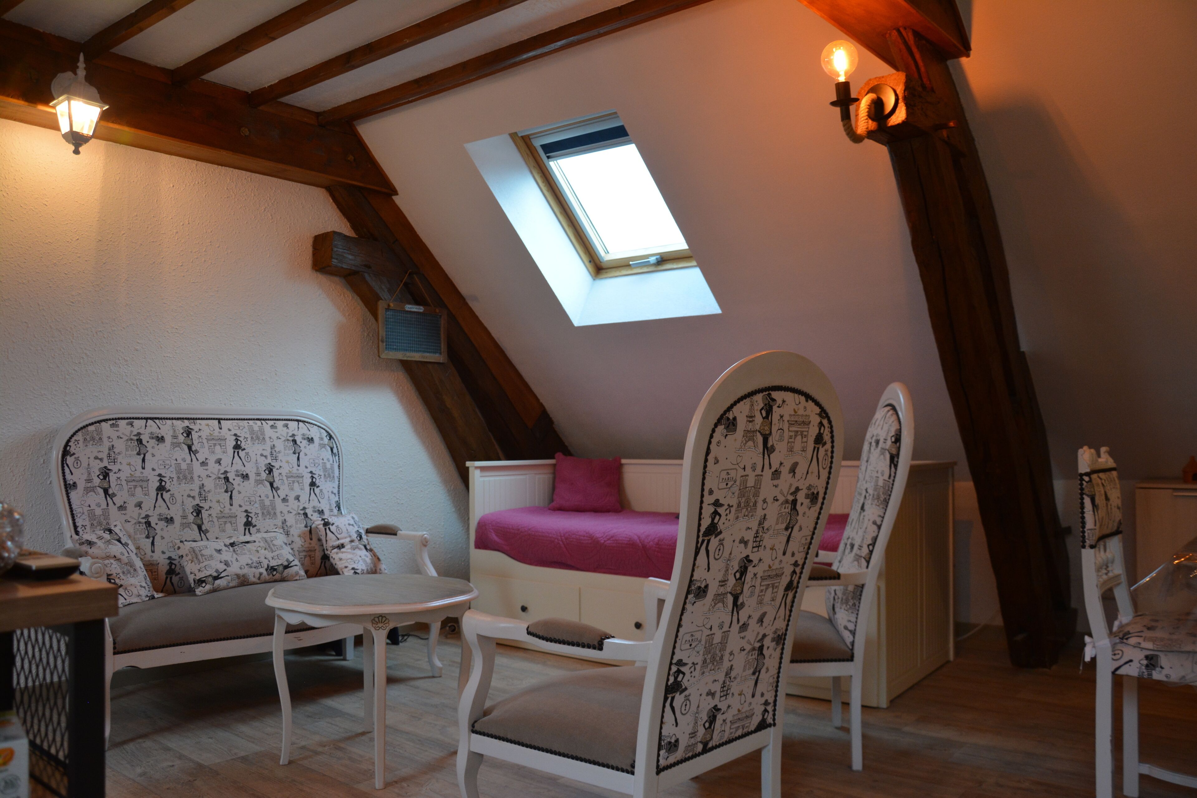 A cozy attic room with a slanted ceiling, a small window, a sofa, two chairs, a table, and a bed with a pink blanket.