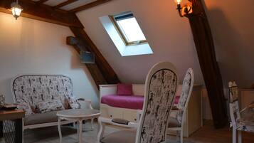 A cozy attic room with a slanted ceiling, a small window, a sofa, two chairs, a table, and a bed with a pink blanket.