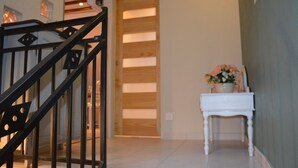 Una escalera con barandillas negras que conduce a una puerta de madera. En el fondo, una mesa blanca con un arreglo floral es visible.