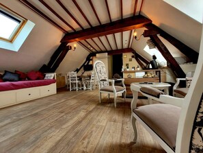 A cozy attic bedroom with a sloped ceiling, wooden beams, a bed with red pillows, a dining table with chairs, and a small kitchen area. - Renovated Loft in the Côte des Blancs near Epernay, the capital of Champagne (Avize)