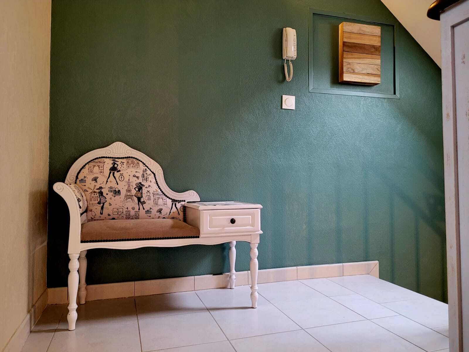 A corner room with a green wall, a white upholstered bench with a patterned cushion, a small white bedside table, and a wall-mounted telephone.