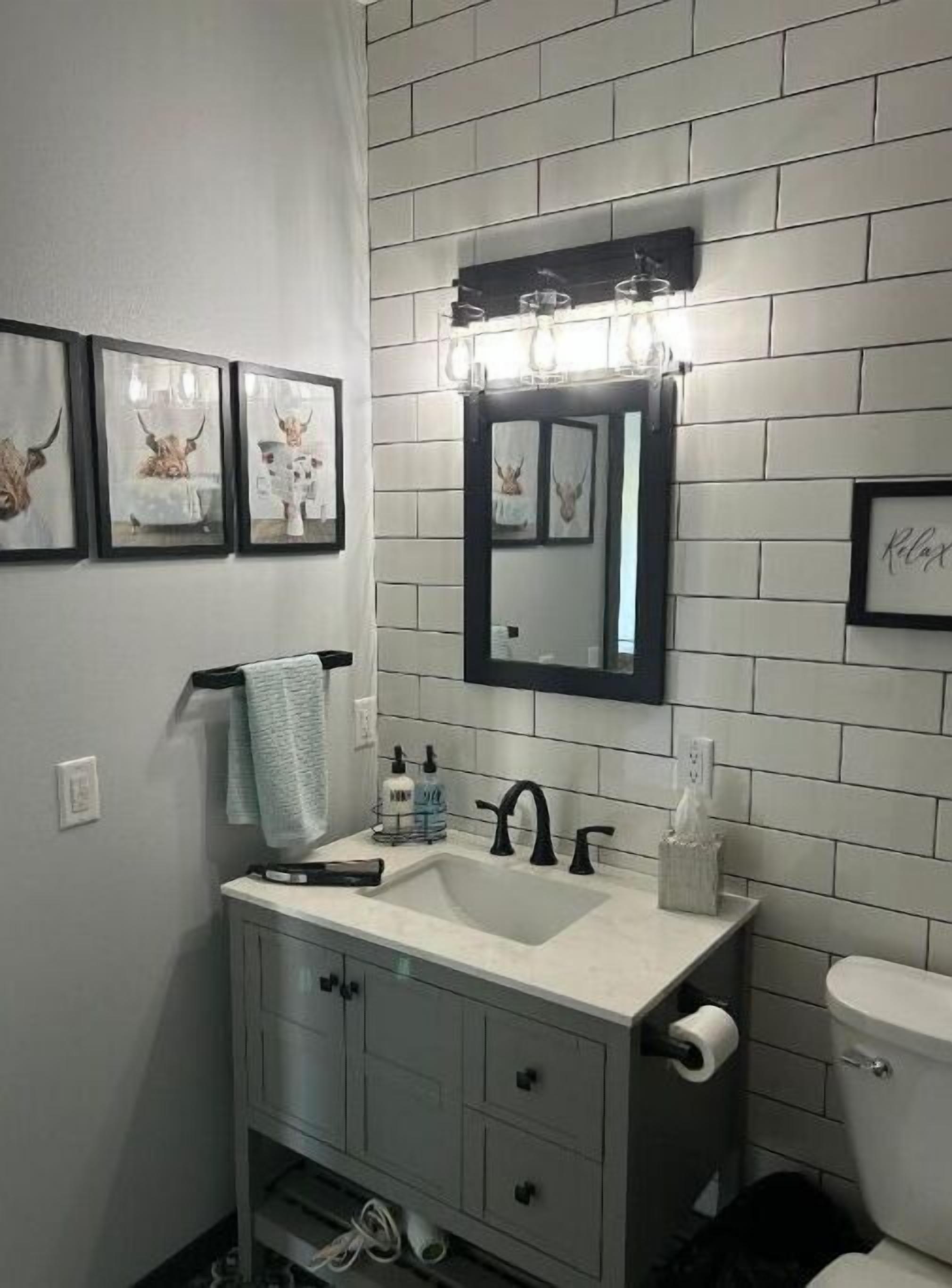 Une salle de bain avec un mur recouvert de carreaux blancs, un lavabo doté de robinets noirs, un miroir et des œuvres d’art encadrées accrochées au mur.