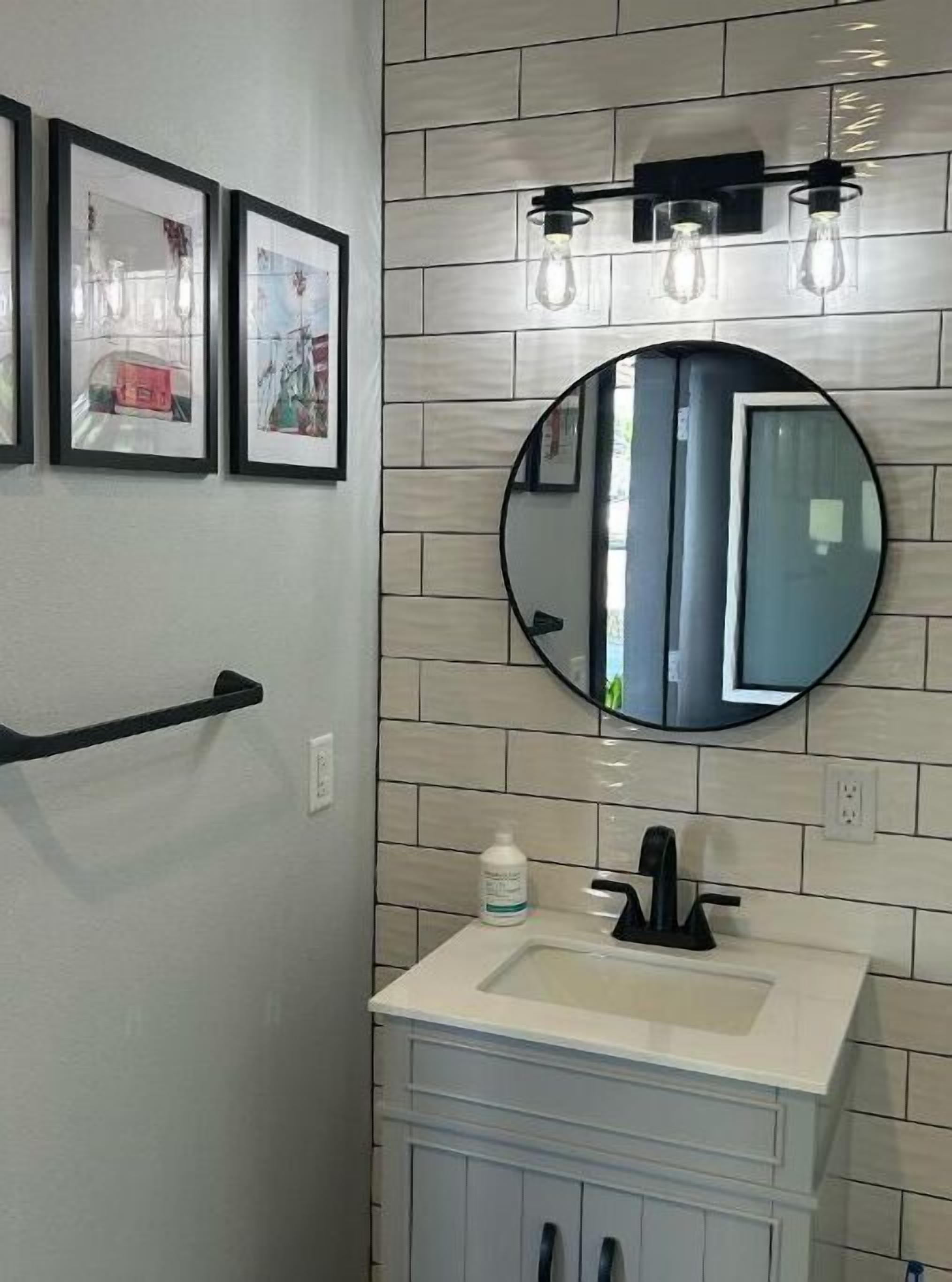 Une salle de bain avec un lavabo blanc, un miroir rond et trois luminaires placés au-dessus du miroir.