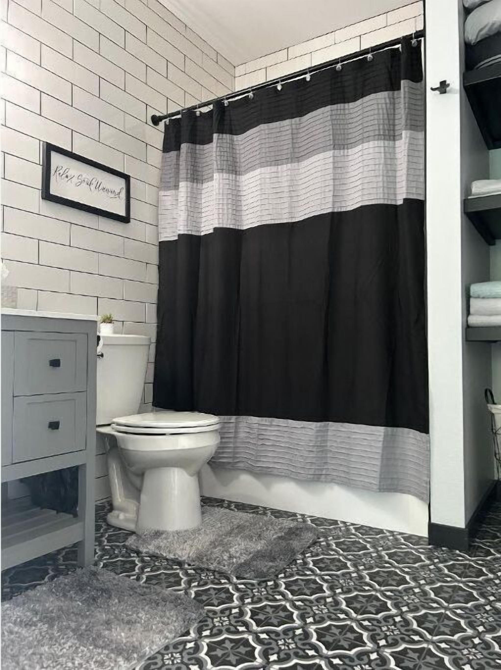 Un baño con pared de azulejos blancos, cortina de ducha a rayas negras y blancas, un inodoro blanco y un tapete estampado.