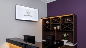 A hotel reception desk with a modern design, a mounted television displaying the hotel name, and a wooden shelving unit with various items. - Herzl Hotel bei Erding (Forstern)