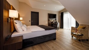 A modern hotel room with a large bed, a desk, and a chair. There is a TV mounted on the wall and a window with curtains. - Herzl Hotel bei Erding (Forstern)