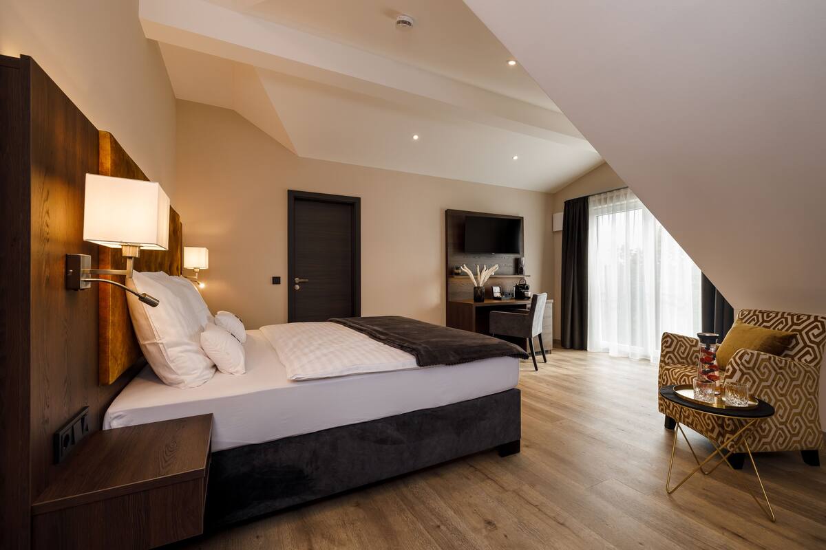 A modern hotel room with a large bed, a desk, and a chair. There is a TV mounted on the wall and a window with curtains.