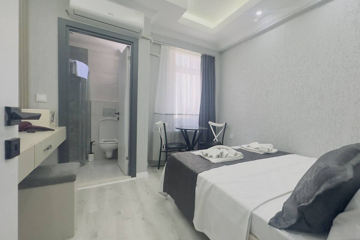 A hotel room with a bed, a desk, a chair, a small table, and a bathroom visible through an open door.