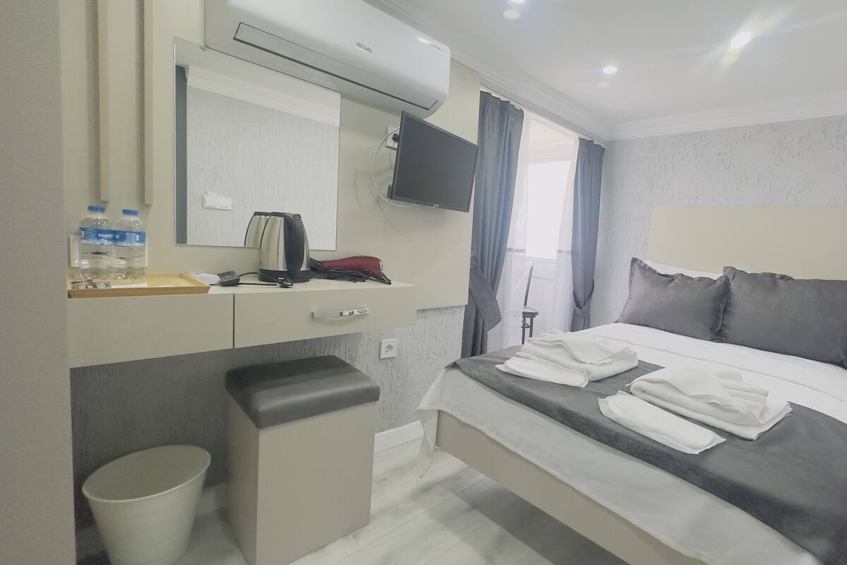 A hotel room with a bed, a desk with a mirror, a chair, a trash bin, and a TV mounted on the wall.