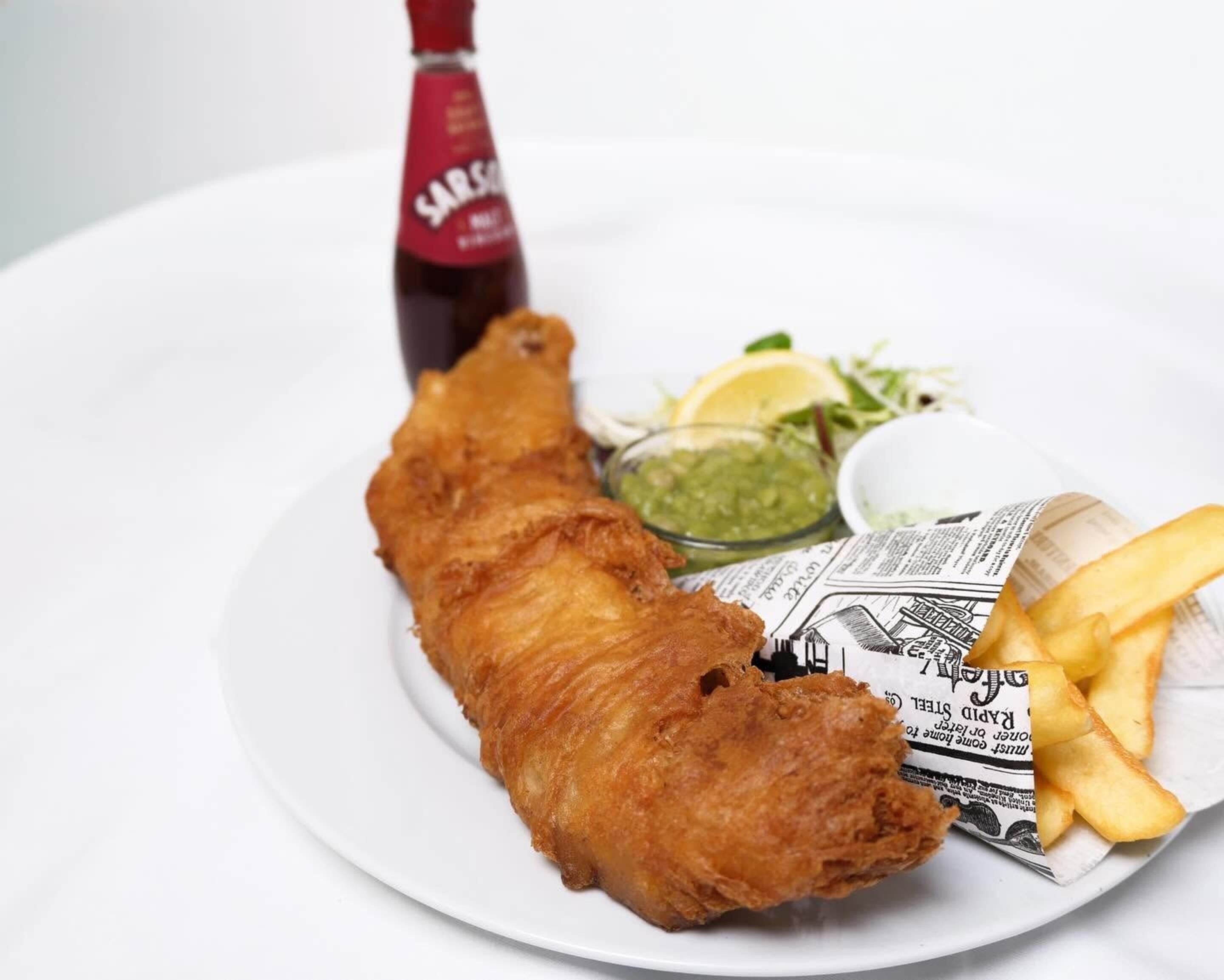 Un plato con un pescado frito grande, gajos de limón, patatas fritas y una botella de salsa.