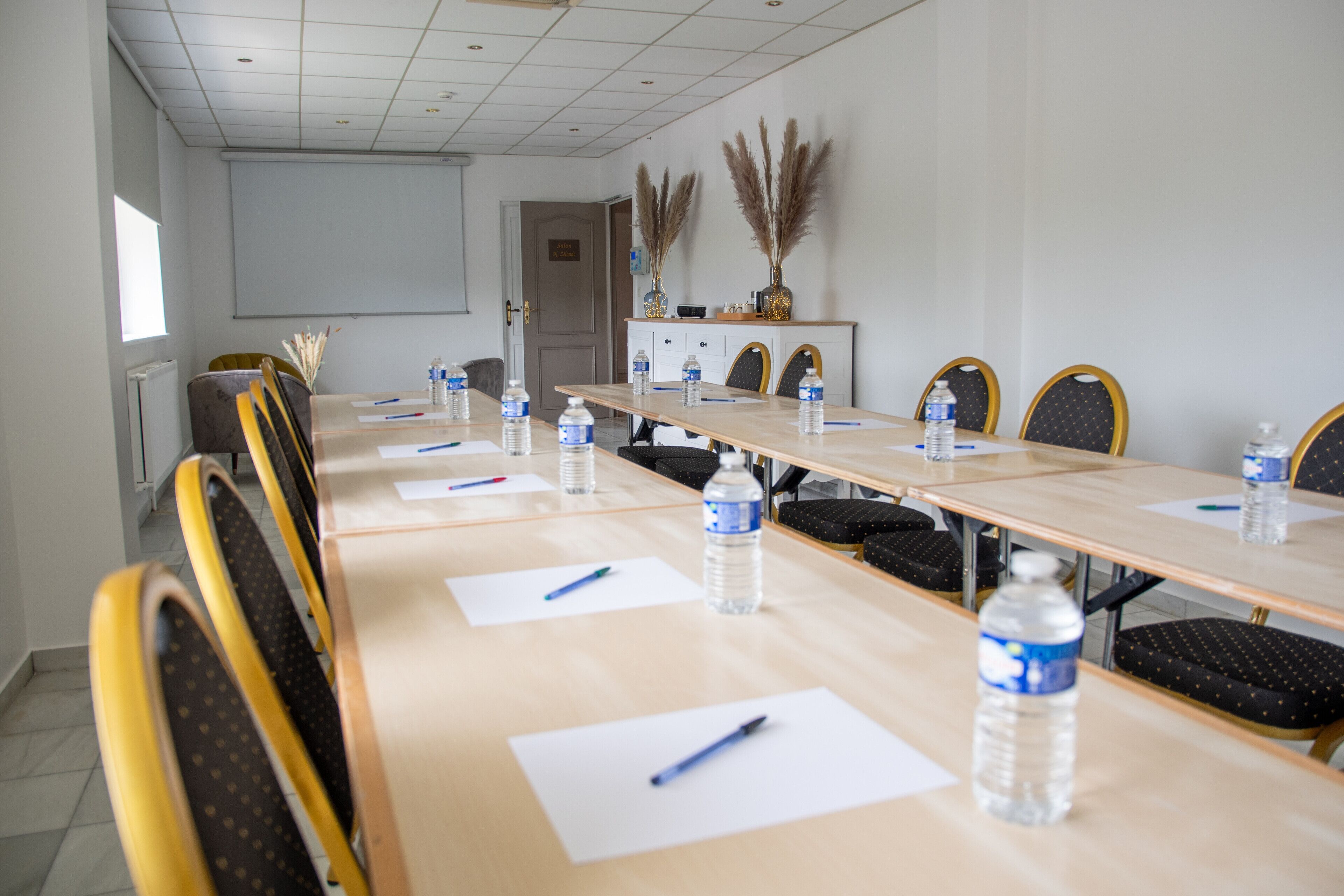 A conference room with a long table, chairs, water bottles, pens, and a projector screen.
