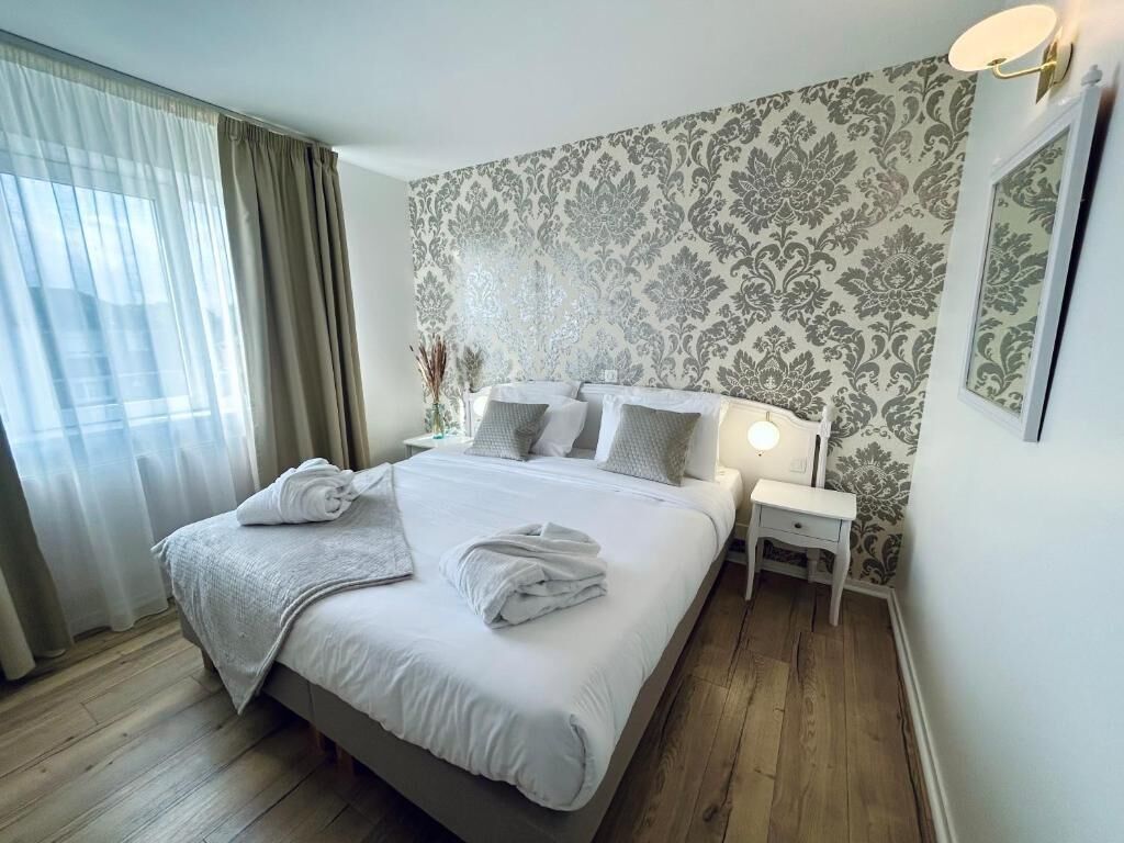 Una cama bien hecha con almohadas y una colcha, una mesita de noche con lámpara, y un papel tapiz estampado en la pared.