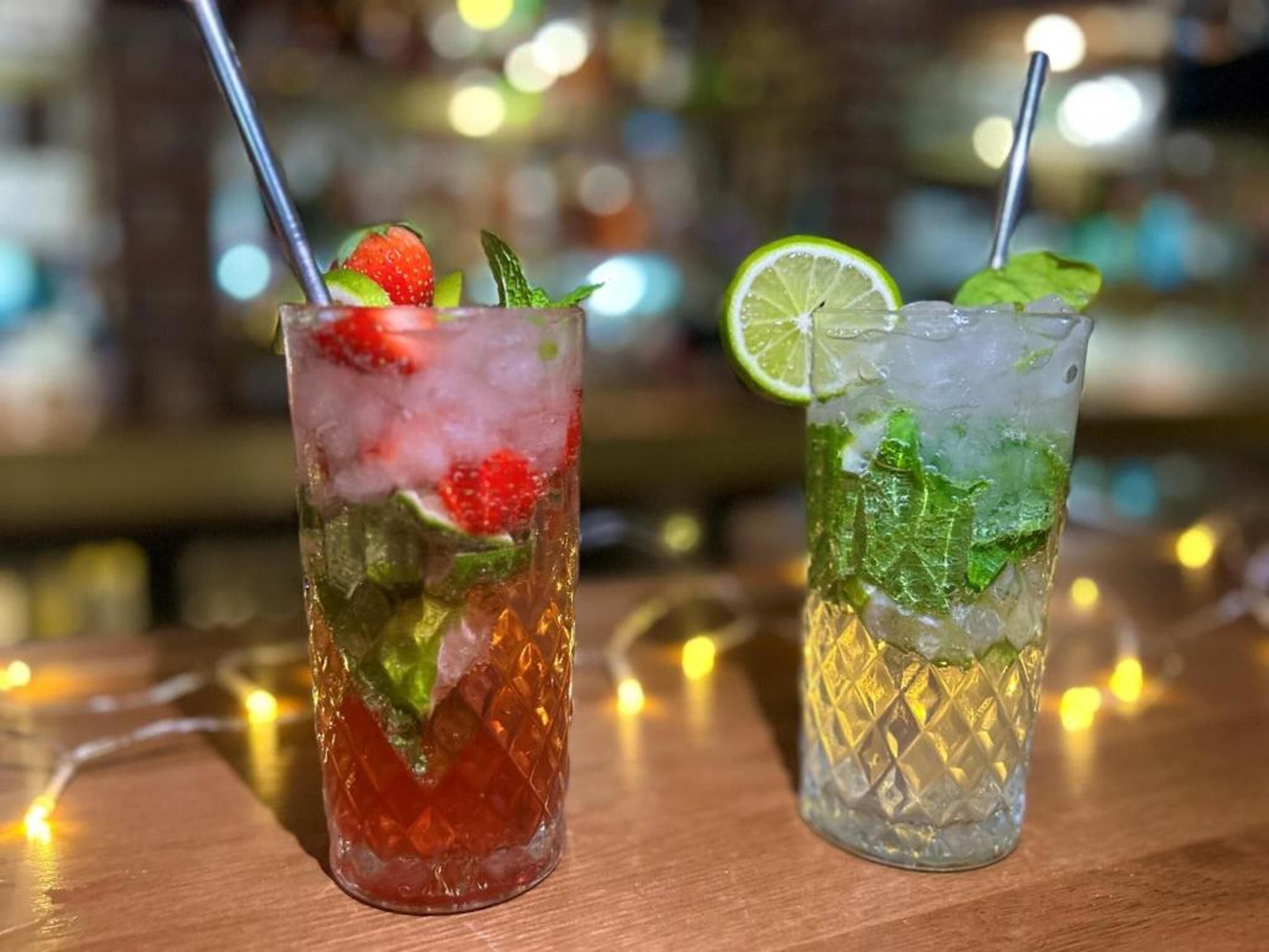 Two glasses of iced drinks with straws, garnished with lime and mint, on a wooden surface with blurred background lights.