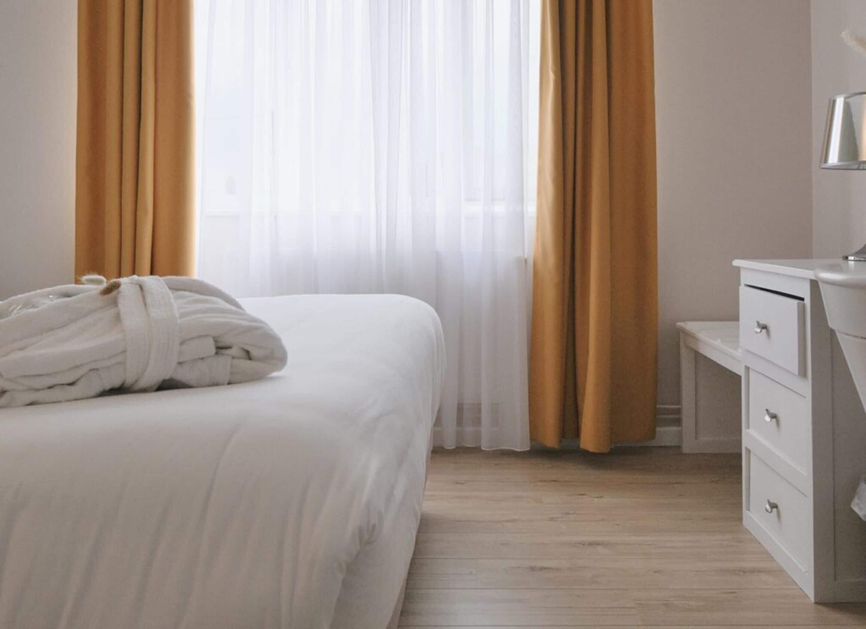 Habitación de hotel con cama, mesita de noche con cajones y ventana con cortinas translúcidas.