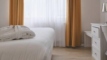 Habitación de hotel con cama, mesita de noche con cajones y ventana con cortinas translúcidas.