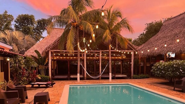 A pool area with a thatched-roof structure, lounge chairs, and string lights. - Hotel Ingigante (Tola)