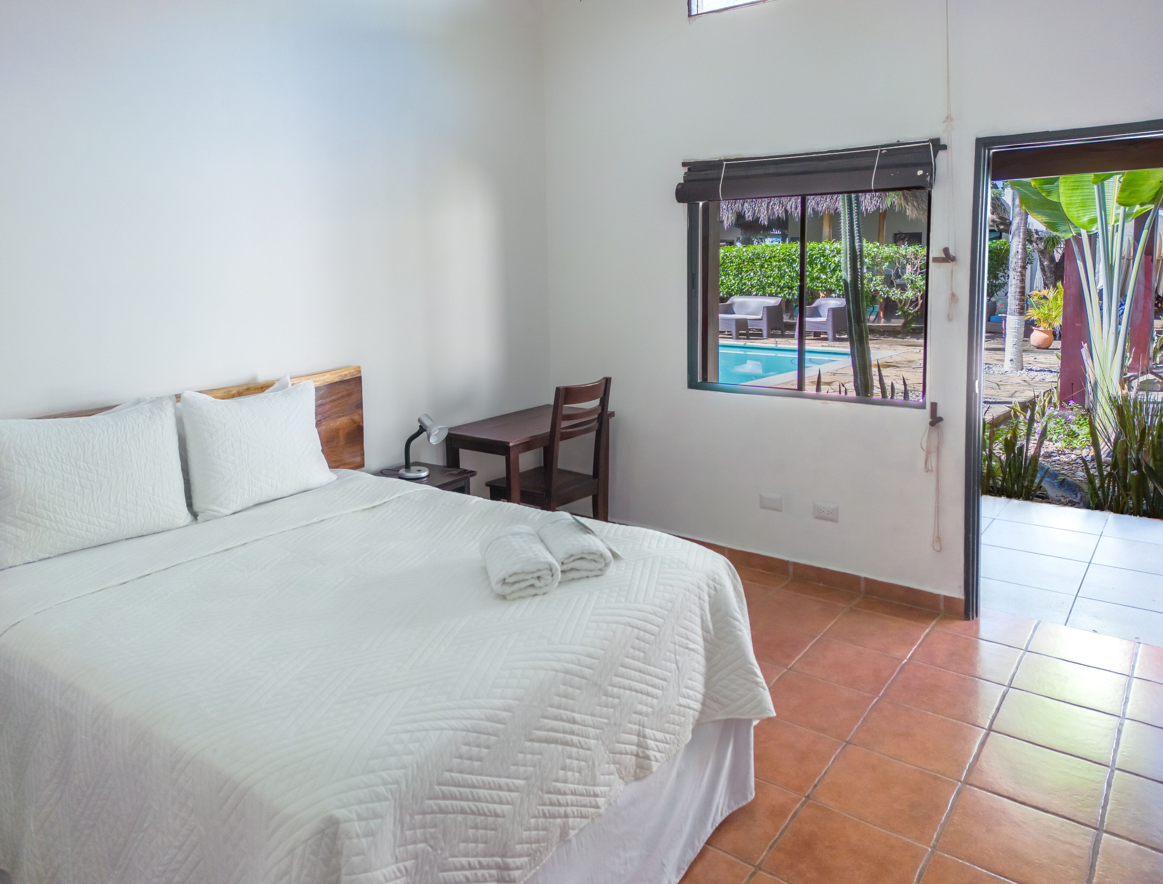 A hotel room with a bed, a desk, a chair, a window with a view of a pool and greenery, and a door.