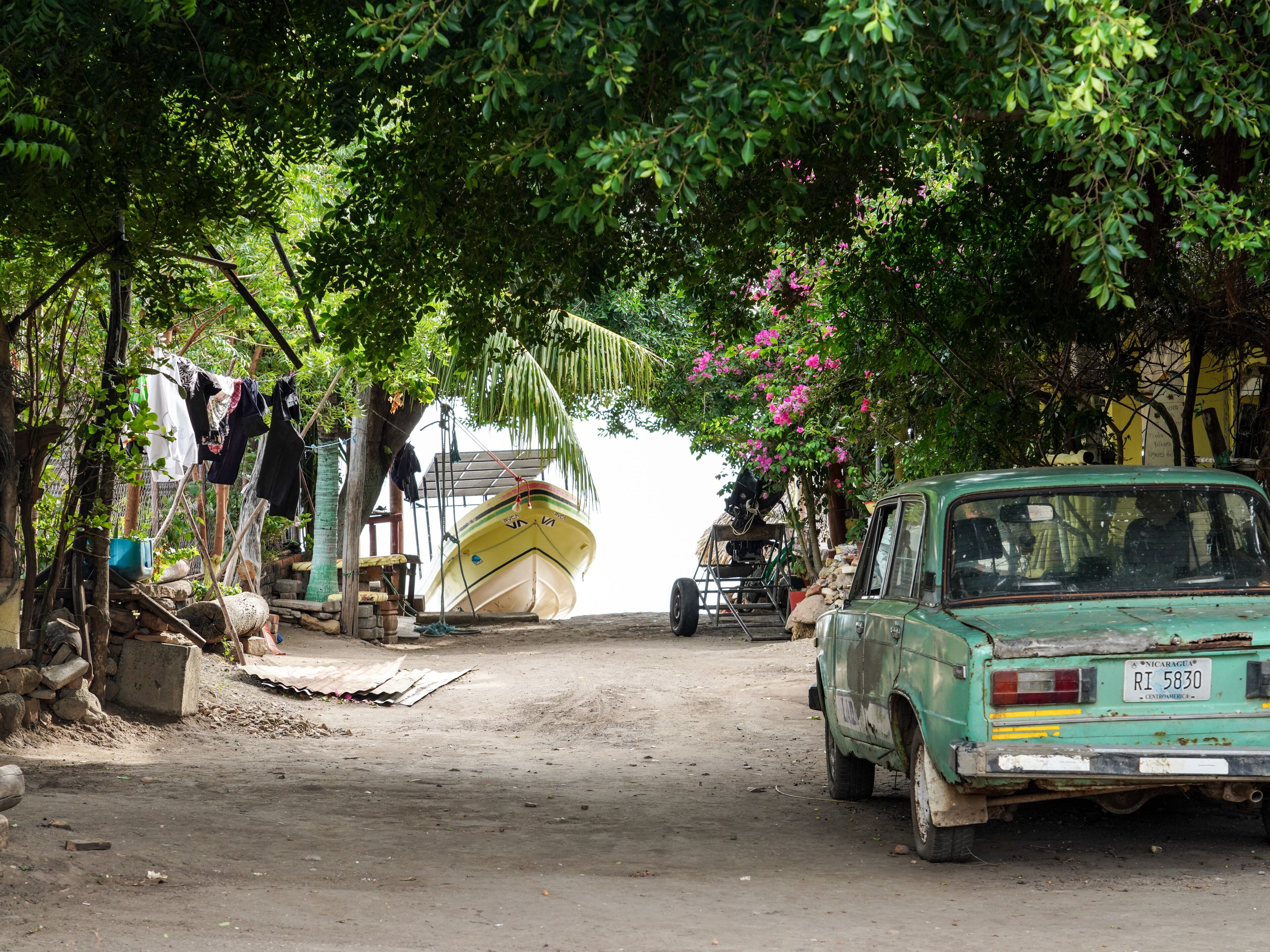 A rustic street scene with a vintage car parked on the side, a boat tied to a tree, and a makeshift clothesline.
