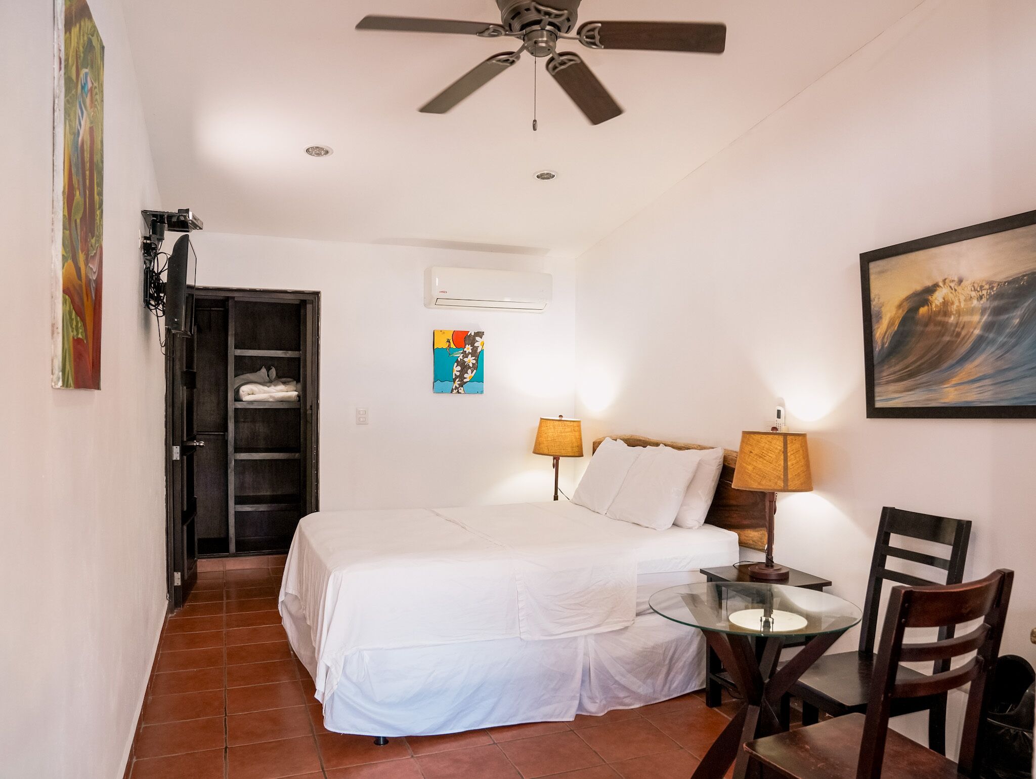 A hotel room with a bed, a desk with a chair, a ceiling fan, and artwork on the walls.