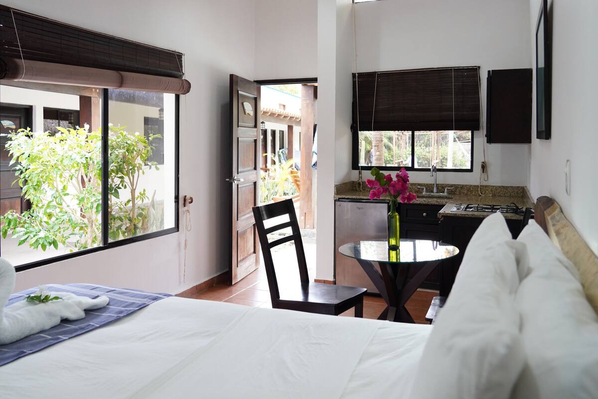 Hotelkamer met een bed, een eettafel met stoelen, een kitchenette en uitzicht op groen.