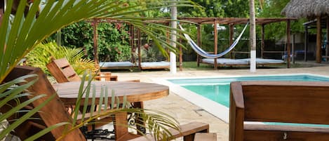 A wooden deck with lounge chairs and a hammock by a pool, surrounded by tropical plants and a thatched-roof structure.