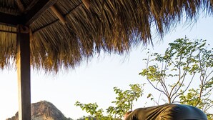 A wooden deck with a thatched roof, a person performing a yoga pose, and a view of a body of water and hills in the background. - Hotel Ingigante (Tola)