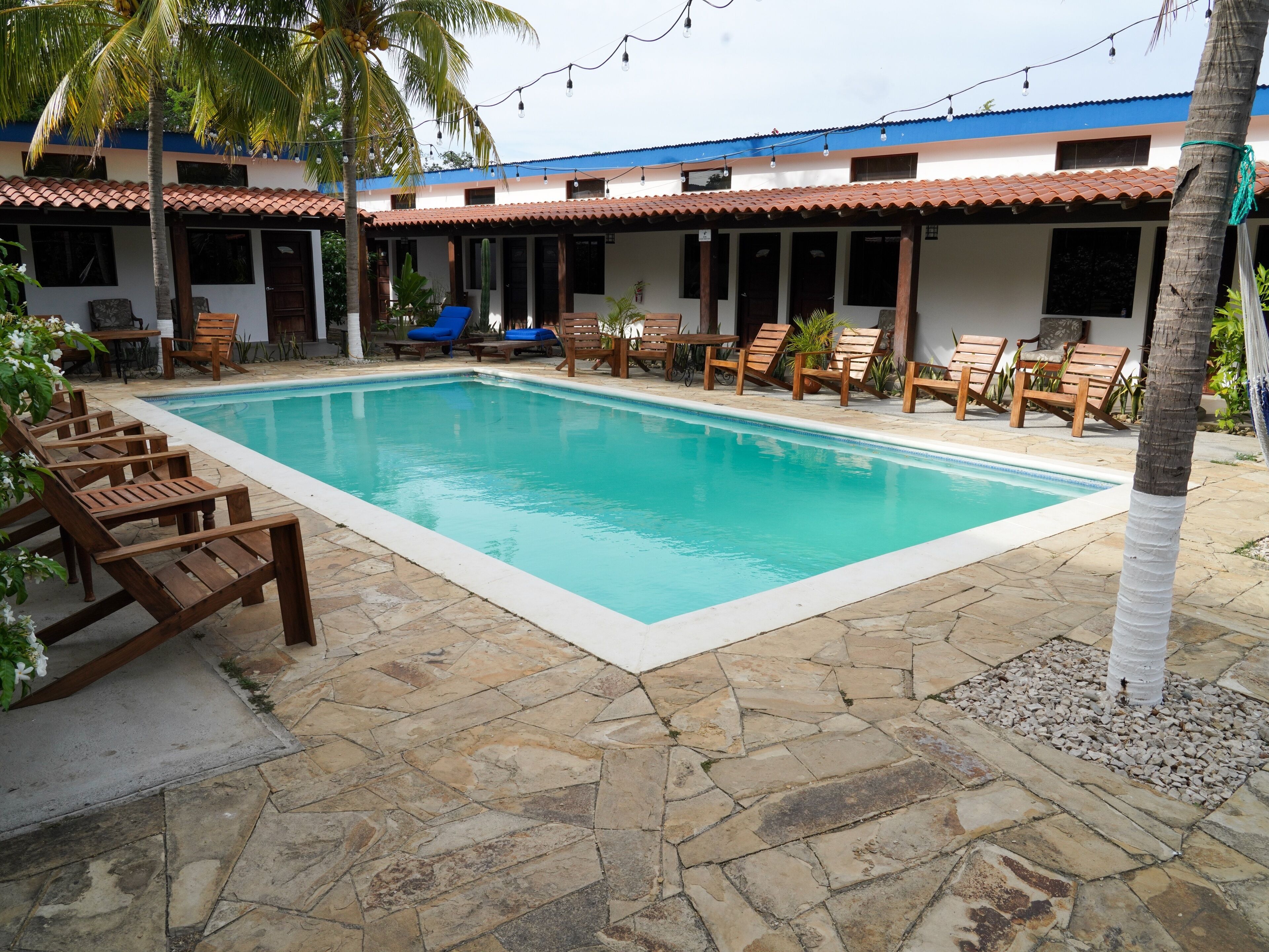 A rectangular pool with a stone tiled deck, surrounded by wooden lounge chairs and a covered seating area with a blue roof.
