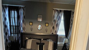Ein Schlafzimmer mit Bett, Kopfteil, Nachttischen, Lampen, Vorhängen und einem an der Wand befestigten Spiegel.